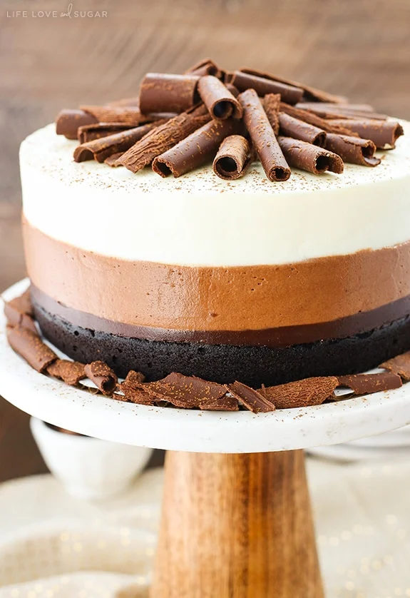 A multi-layered chocolate cake with a dark chocolate base, milk chocolate middle, and white chocolate or cream top, adorned with chocolate curls and a dusting of cocoa powder or powdered sugar, displayed on a white pedestal stand.