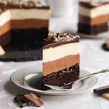 This stunning multi-layered chocolate cake features a rich dark chocolate base, a creamy milk chocolate middle layer, and a smooth white chocolate or cream top layer. Decorated with elegant chocolate curls and a light dusting of cocoa powder or powdered sugar, this dessert is beautifully presented on a white pedestal stand.
